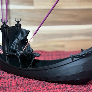 Könnte beinhalten: Ein schwarzer Räucherstäbchenhalter in Form einer Gondel mit einer Kapuzenfigur in der Mitte. Der Halter hat ein dekoratives Design und hält lila Räucherstäbchen. Die Gondel steht auf einer rot gemusterten Oberfläche.