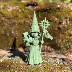 Puede incluir: Una figura en miniatura de un mago impresa en 3D de color verde, que lleva un sombrero puntiagudo y sostiene un bastón con una punta en forma de estrella. El mago lleva una túnica larga y una mochila.