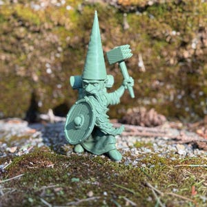 Puede incluir: Una figurita de plástico verde de un gnomo con un sombrero puntiagudo, sosteniendo un escudo y un martillo. El gnomo tiene barba y está de pie sobre un lecho de musgo.