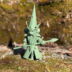 Puede incluir: Una figurita de plástico verde de un gnomo con un sombrero puntiagudo y sosteniendo una lanza. El gnomo tiene una larga barba blanca y lleva una túnica con detalles intrincados.