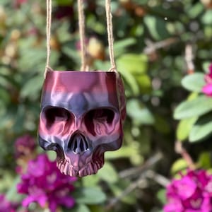 May include: A hanging planter shaped like a skull, made of a dark red and black iridescent material. The planter is hanging from a natural twine cord.