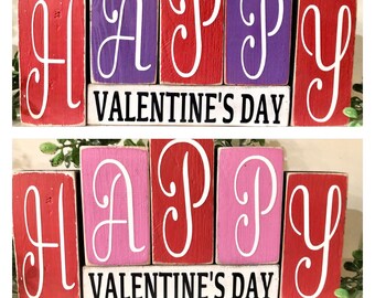 May include: Wooden block letters spelling "HAPPY VALENTINE'S DAY" in red, purple, and pink with white lettering. The blocks are arranged in two rows, with the word "HAPPY" on top and "VALENTINE'S DAY" on the bottom.