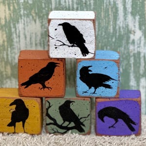 May include: A stack of seven square wooden blocks, each painted with a different color and a black raven silhouette. The blocks are painted in various colors, including white, orange, blue, yellow, green, and purple.