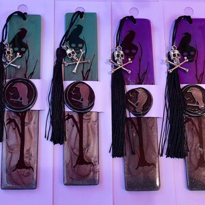 May include: Set of ten bookmarks featuring a black silhouette of a tree with a crow perched on a branch. Each bookmark has a different colored background, including red, orange, green, purple, and brown. Each bookmark has a black tassel and a silver skull and crossbones charm.