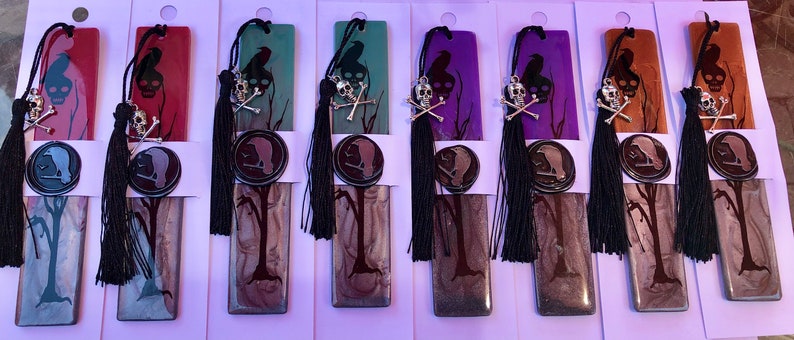 May include: Set of ten bookmarks featuring a black silhouette of a tree with a crow perched on a branch. Each bookmark has a different colored background, including red, orange, green, purple, and brown. Each bookmark has a black tassel and a silver skull and crossbones charm.
