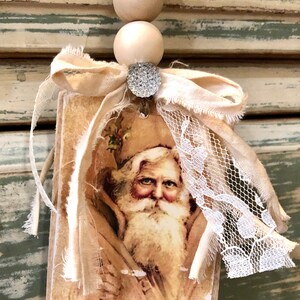 May include: A vintage-style Christmas ornament featuring a portrait of Father Christmas on a wooden tag with a beige fabric bow and lace trim. The ornament is adorned with a sparkly rhinestone.