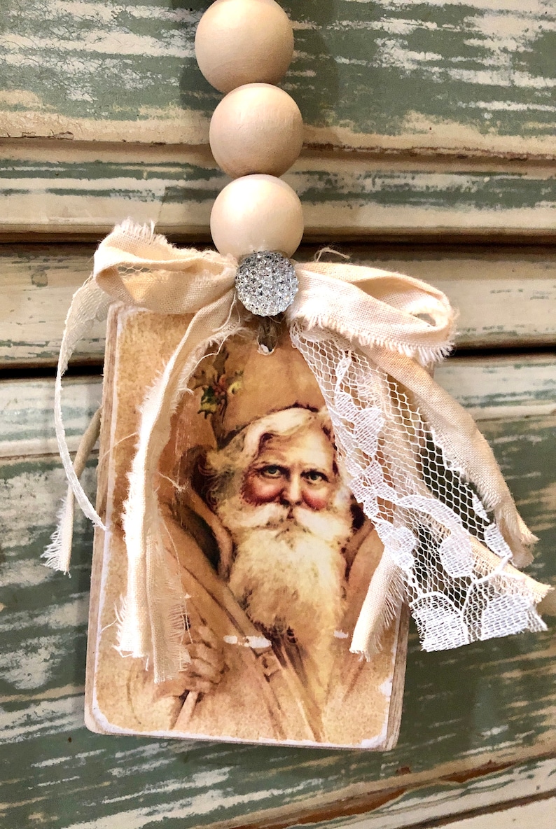 May include: A vintage-style Christmas ornament featuring a portrait of Father Christmas on a wooden tag with a beige fabric bow and lace trim. The ornament is adorned with a sparkly rhinestone.