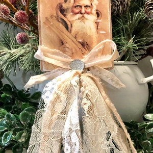 May include: A vintage-style Christmas ornament featuring a neutral-toned image of Santa Claus on a wooden card with a white lace bow and pearl accents. The ornament is surrounded by greenery.