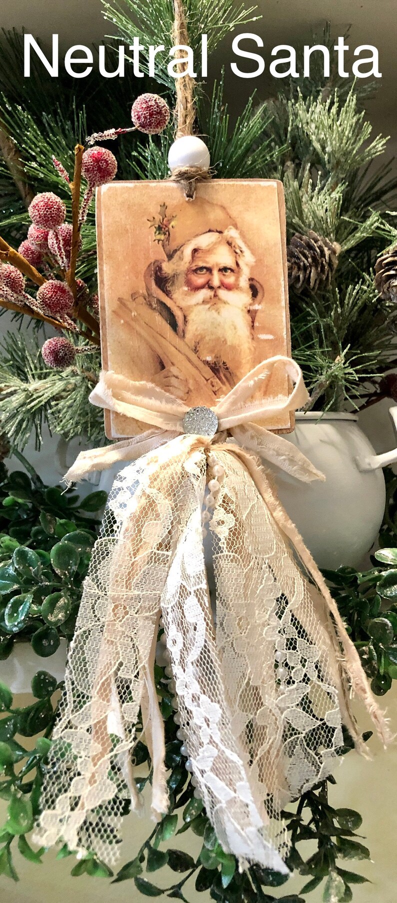 May include: A vintage-style Christmas ornament featuring a neutral-toned image of Santa Claus on a wooden card with a white lace bow and pearl accents. The ornament is surrounded by greenery.