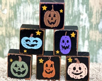 May include: A stack of seven black wooden blocks with various colored jack-o'-lantern designs. Each block features a different pumpkin color, including brown, blue, purple, green, orange, and white, with yellow star accents. The blocks are arranged in a pyramid shape.
