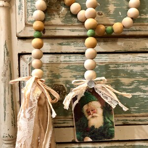 May include: A wooden bead garland with green and beige beads, finished with a fabric tassel and a vintage Santa Claus image.