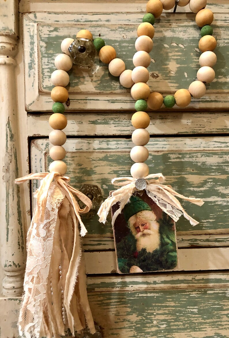 May include: A wooden bead garland with green and beige beads, finished with a fabric tassel and a vintage Santa Claus image.