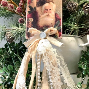 May include: A vintage Christmas card ornament featuring a red-suited Santa Claus with a white beard and a red hat. The card is decorated with a white lace trim and a white bow with a sparkly rhinestone center. The ornament is hanging from a brown twine string.