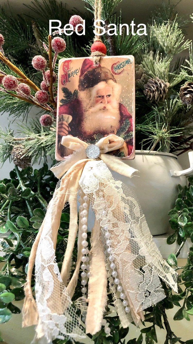 May include: A vintage Christmas card ornament featuring a red-suited Santa Claus with a white beard and a red hat. The card is decorated with a white lace trim and a white bow with a sparkly rhinestone center. The ornament is hanging from a brown twine string.