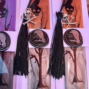 May include: Three handmade bookmarks with a black raven, skull and crossbones, and a tree design. The bookmarks are made of resin and have a silver and black color scheme. Each bookmark has a black tassel.