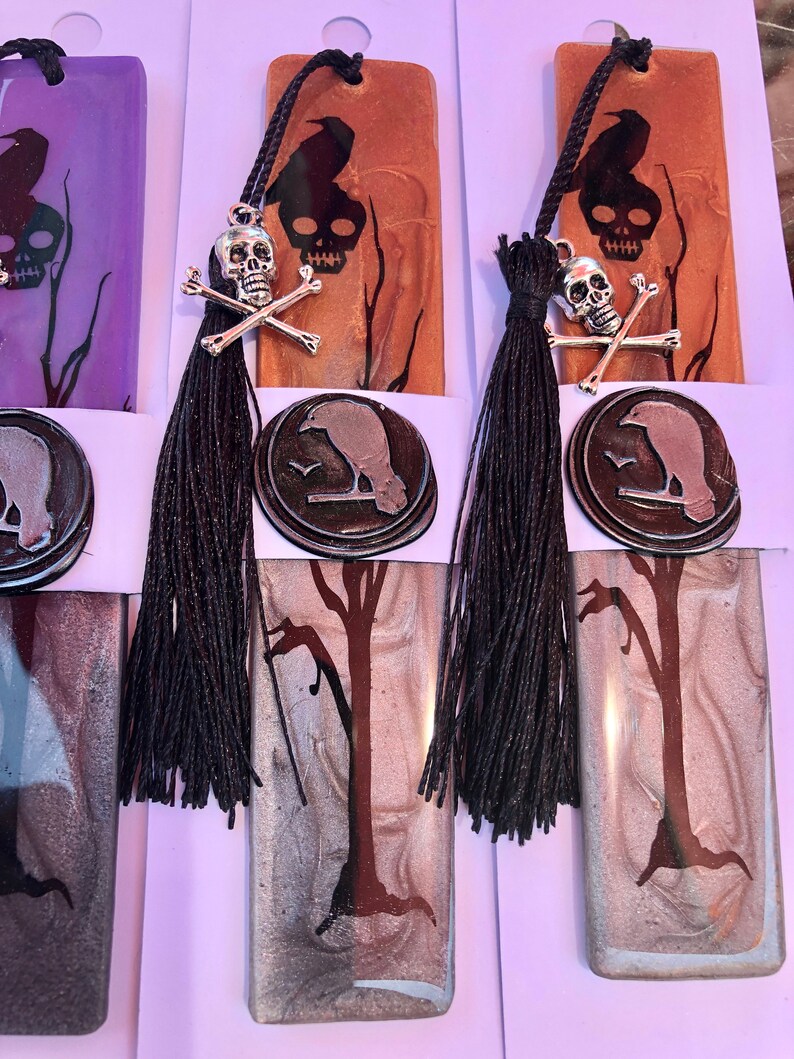 May include: Three handmade bookmarks with a black raven, skull and crossbones, and a tree design. The bookmarks are made of resin and have a silver and black color scheme. Each bookmark has a black tassel.