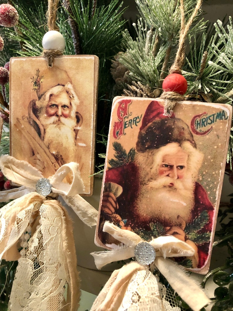 May include: Two vintage Christmas ornaments featuring images of Santa Claus. Each ornament is made of wood and has a fabric bow with a sparkly embellishment. The ornaments are hung with twine and have a white and red bead at the top.