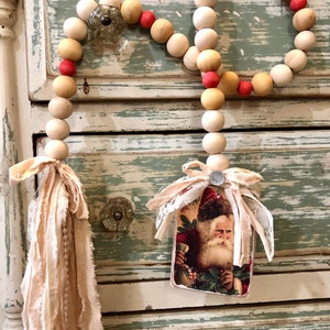 May include: A wooden bead garland with red accents and a vintage Santa Claus image attached. The garland is hanging on a distressed white and green cabinet.
