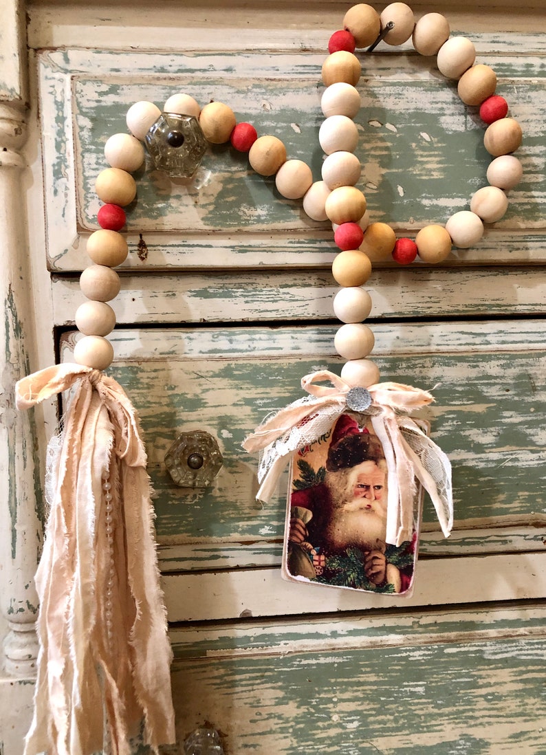 May include: A wooden bead garland with red accents and a vintage Santa Claus image attached. The garland is hanging on a distressed white and green cabinet.