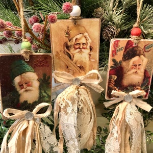 May include: Three vintage Christmas ornaments featuring Santa Claus images. Each ornament is a rectangular card with a lace trim and a bow. The cards are attached to a twine string with a wooden bead.