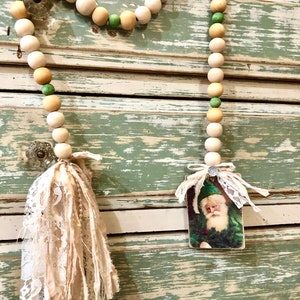 May include: A white and green beaded garland with a vintage Santa Claus image and two white fabric tassels. The garland is hanging on a distressed white and green painted wooden surface.