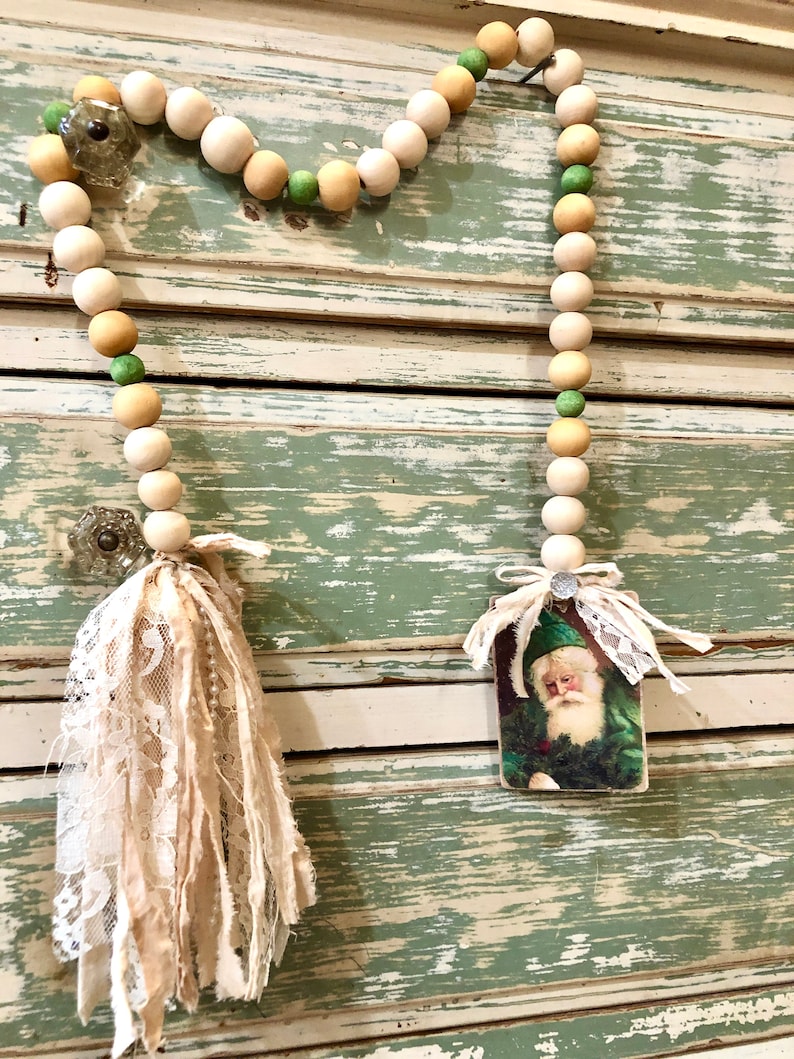 May include: A white and green beaded garland with a vintage Santa Claus image and two white fabric tassels. The garland is hanging on a distressed white and green painted wooden surface.