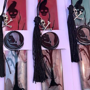 May include: Three bookmarks with a black raven and skull design. The bookmarks are made of resin and have a silver and black marbled effect. Each bookmark has a black tassel.