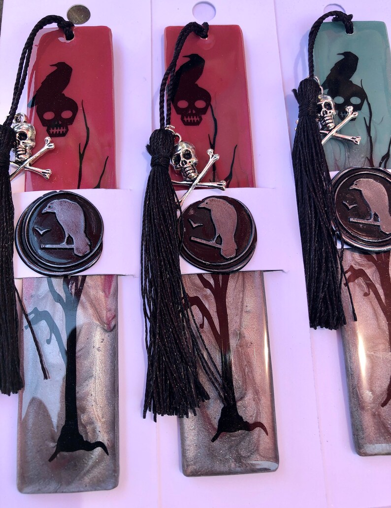 May include: Three bookmarks with a black raven and skull design. The bookmarks are made of resin and have a silver and black marbled effect. Each bookmark has a black tassel.