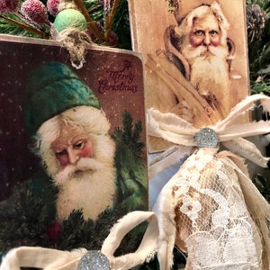 May include: Two vintage Christmas ornaments featuring illustrations of Santa Claus. Each ornament is decorated with a white lace bow and a sparkly silver button. The ornaments are hanging from a green pine branch.