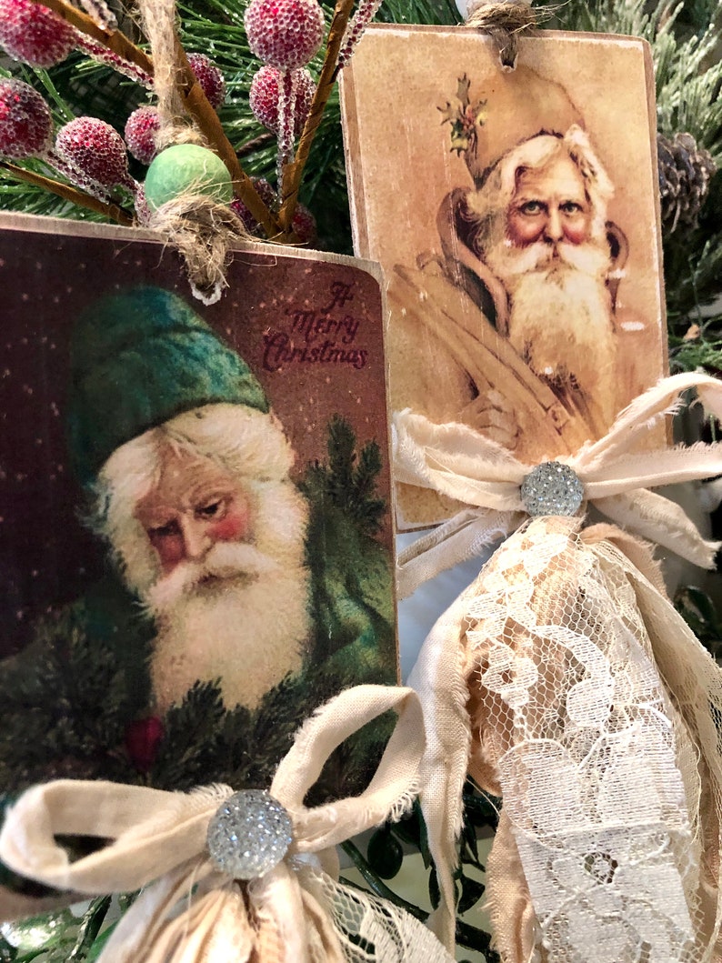 May include: Two vintage Christmas ornaments featuring illustrations of Santa Claus. Each ornament is decorated with a white lace bow and a sparkly silver button. The ornaments are hanging from a green pine branch.