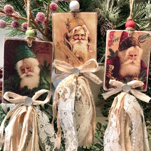 May include: Three vintage-style Christmas ornaments featuring images of Santa Claus. Each ornament is made of wood and has a white lace trim and a brown ribbon bow. The ornaments are hanging from a branch of a Christmas tree.