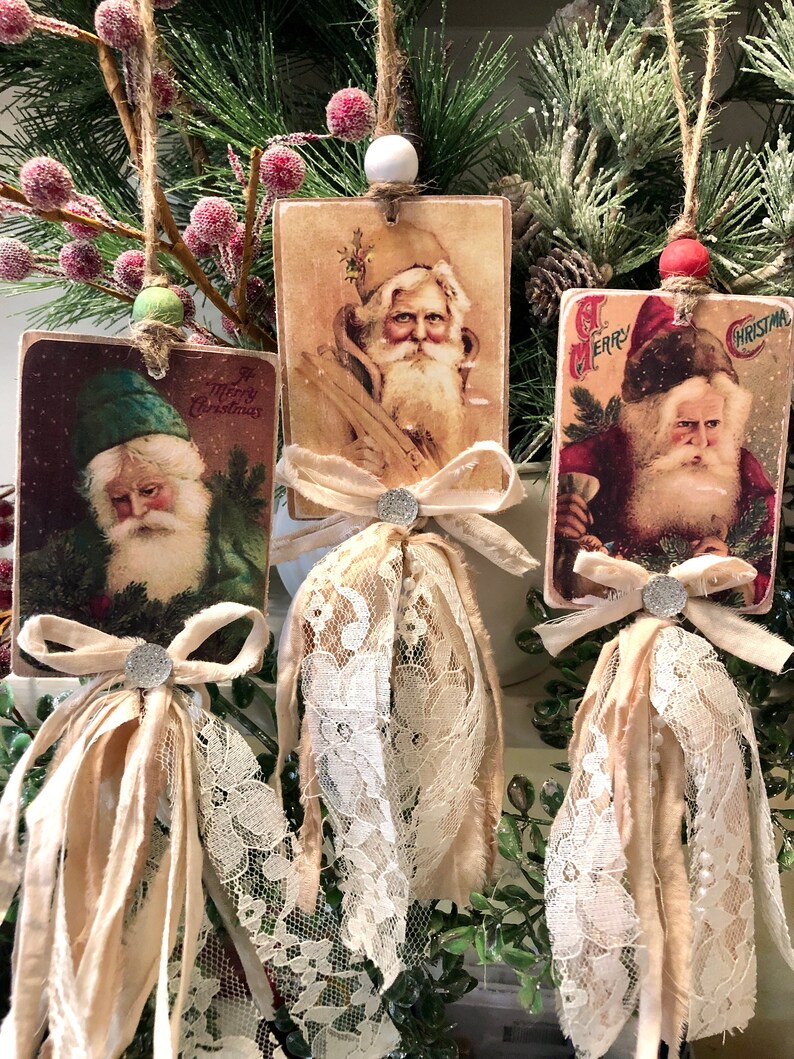 May include: Three vintage-style Christmas ornaments featuring images of Santa Claus. Each ornament is made of wood and has a white lace trim and a brown ribbon bow. The ornaments are hanging from a branch of a Christmas tree.