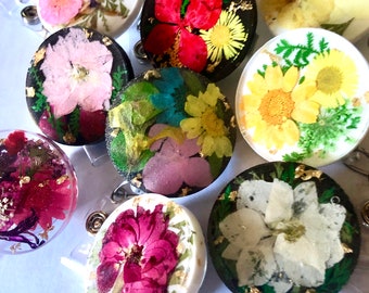 May include: A selection of round badge reels, each encased in resin and adorned with pressed flowers. The reels showcase diverse floral arrangements, including red, pink, yellow, and white blooms, accented with gold leaf. Each reel has a clear plastic clip on the reverse.