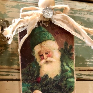 May include: A vintage Christmas bauble featuring a Santa Claus illustration with a green hat and beard, holding a sprig of pine. The bauble is made of wood and has a white ribbon and lace trim. The text "Merry Christmas" is visible on the bauble.