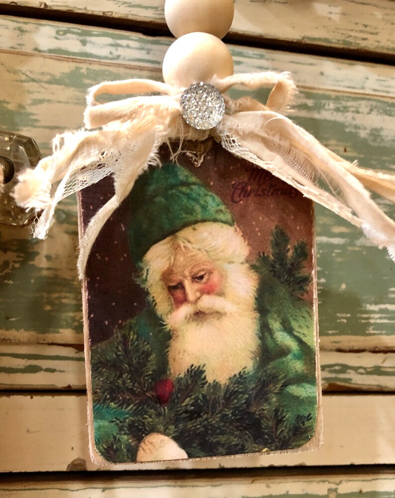 May include: A vintage Christmas bauble featuring a Santa Claus illustration with a green hat and beard, holding a sprig of pine. The bauble is made of wood and has a white ribbon and lace trim. The text "Merry Christmas" is visible on the bauble.