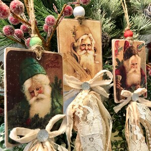 May include: Three vintage-style Christmas ornaments featuring images of Santa Claus. Each ornament is made of wood and has a white lace trim and a brown ribbon bow. The ornaments are hanging from a green branch with red berries.