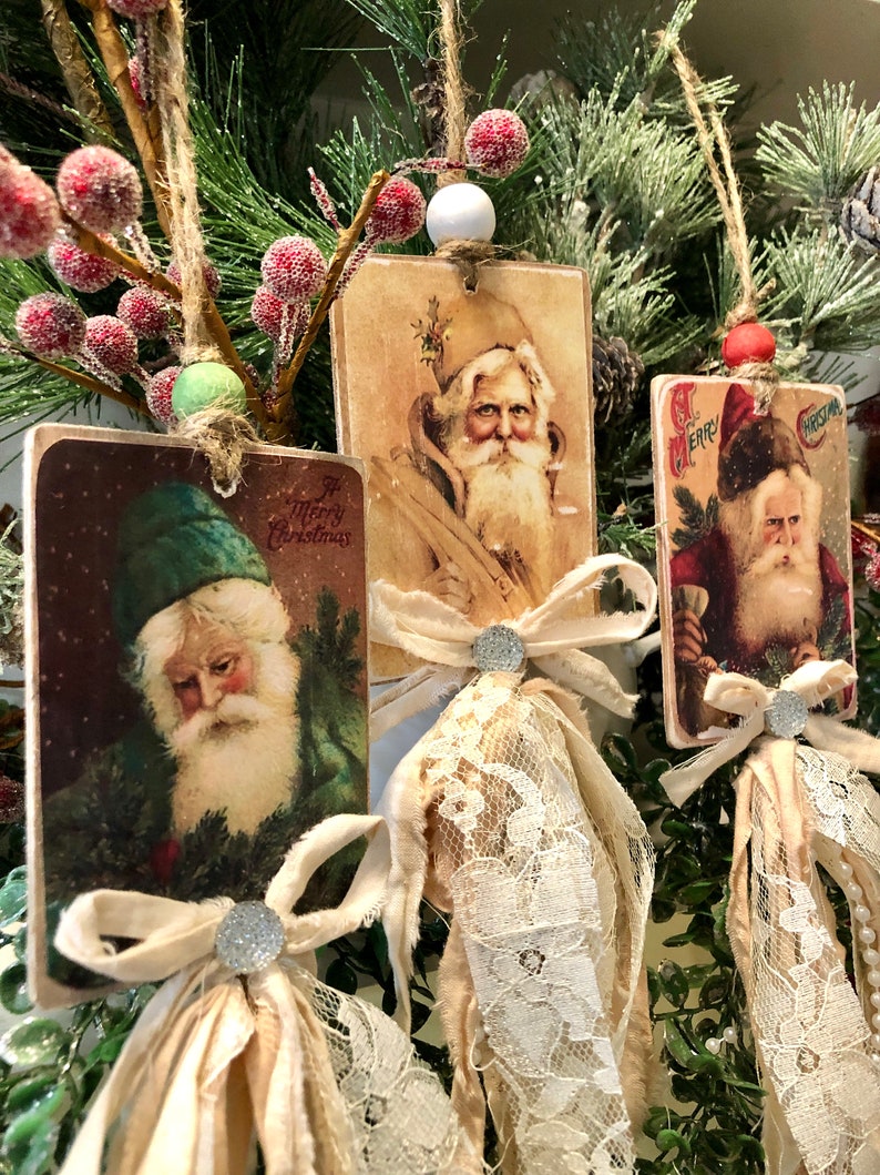 May include: Three vintage-style Christmas ornaments featuring images of Santa Claus. Each ornament is made of wood and has a white lace trim and a brown ribbon bow. The ornaments are hanging from a green branch with red berries.