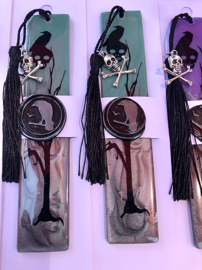 May include: Three black and silver bookmark charms with a raven, skull and crossbones, and a wax seal design. The bookmarks are made of resin and have a black tassel.
