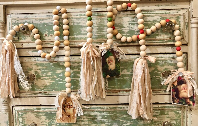 May include: A wooden bead garland featuring vintage Santa Claus images and fabric tassels. The garland is made from natural wood beads with white and red accents. The tassels are made of a soft, beige fabric.