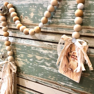 May include: A wooden bead garland with a vintage Santa Claus image attached. The garland is made of natural wood beads in shades of beige and white. The beads are strung together with a white twine. The Santa Claus image is on a card with a white lace trim. The garland is hanging on a distressed wooden surface.