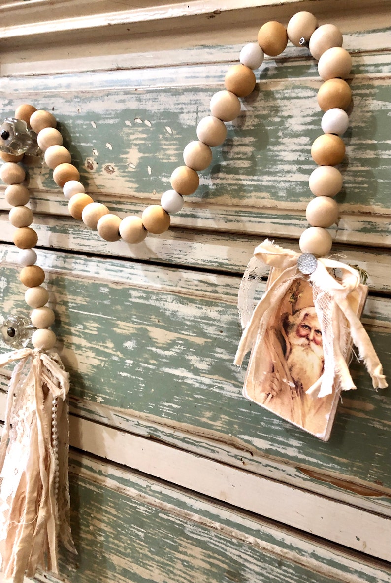 May include: A wooden bead garland with a vintage Santa Claus image attached. The garland is made of natural wood beads in shades of beige and white. The beads are strung together with a white twine. The Santa Claus image is on a card with a white lace trim. The garland is hanging on a distressed wooden surface.