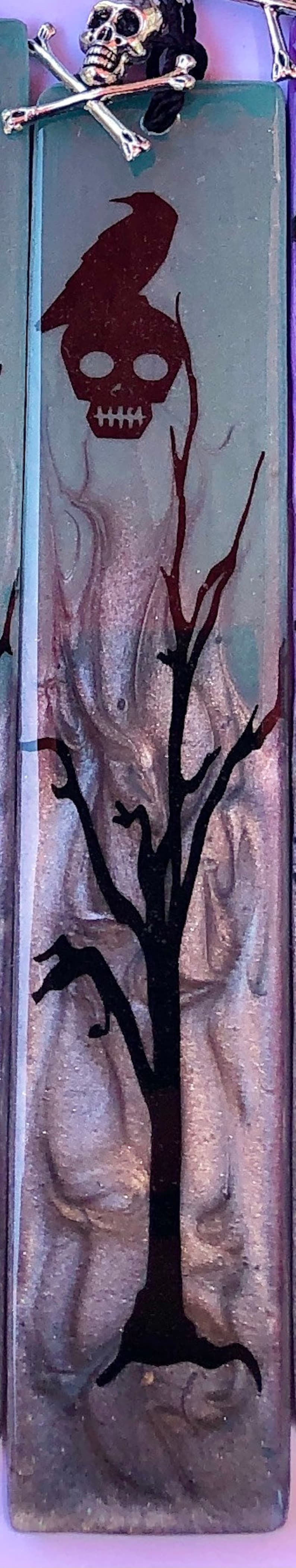 May include: A purple and blue rectangular tile with a black silhouette of a skull with a crow perched on top of it. The skull is surrounded by a black silhouette of a bare tree with branches reaching up.