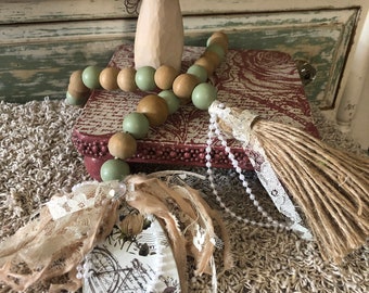 May include: A decorative arrangement featuring a wooden angel figurine holding red objects, a beaded garland with green and brown beads, and a bicycle-themed ornament with lace and ribbon accents. A tassel and a small red box are also present.