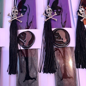 May include: Purple and silver bookmark with a black raven, skull and crossbones, and a black tassel. The bookmark is made of resin and has a glossy finish.