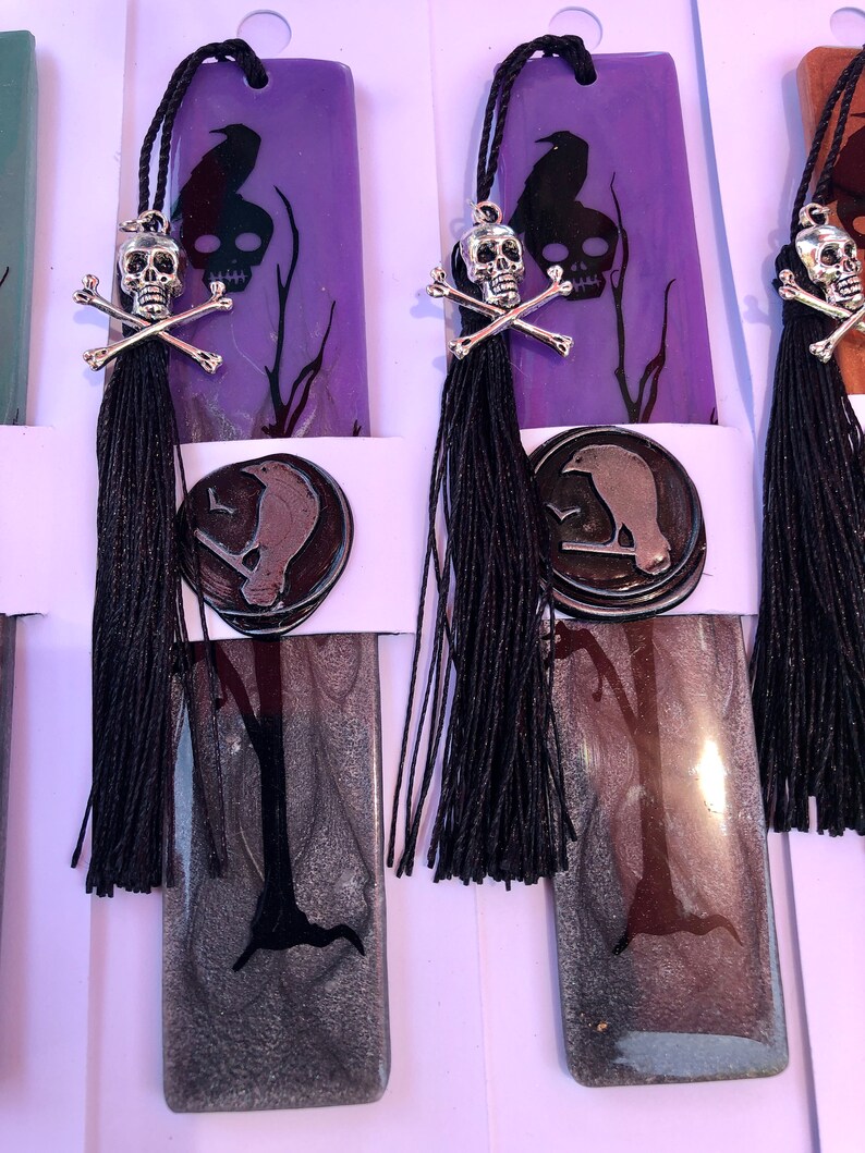 May include: Purple and silver bookmark with a black raven, skull and crossbones, and a black tassel. The bookmark is made of resin and has a glossy finish.
