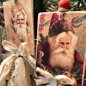 May include: Two vintage Christmas ornaments featuring images of Santa Claus. The ornaments are made of wood and have a rustic, distressed look. Each ornament is adorned with a white lace bow and a silver button.