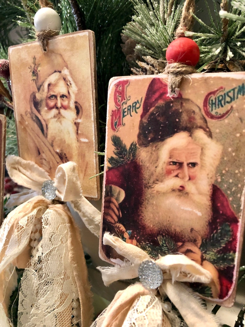 May include: Two vintage Christmas ornaments featuring images of Santa Claus. The ornaments are made of wood and have a rustic, distressed look. Each ornament is adorned with a white lace bow and a silver button.