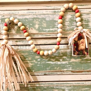 May include: A Christmas garland made of wooden beads in white, beige, and pink with a fabric tassel on one end and a vintage Santa Claus card on the other end.