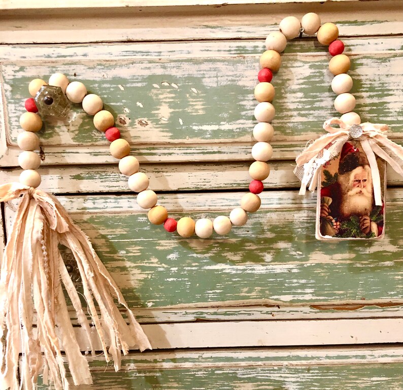 May include: A Christmas garland made of wooden beads in white, beige, and pink with a fabric tassel on one end and a vintage Santa Claus card on the other end.