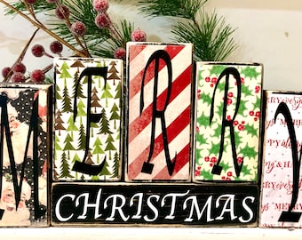 May include: Wooden block sign spelling "Merry Christmas" with different Christmas-themed patterns on each block. The "M" block has a Santa Claus pattern, the "E" block has a green Christmas tree pattern, the "R" block has a red and white candy cane pattern, the "R" block has a green holly pattern, and the "Y" block has a pink and white pattern with the word "Merry" repeated.